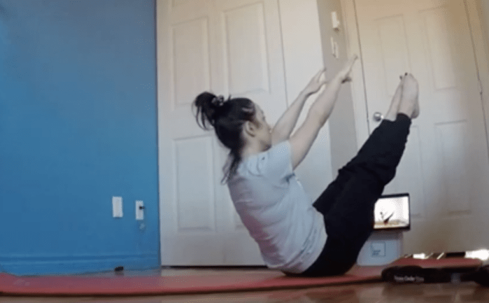 L'instructeur réalise l'exercice Teaser sur un tapis de Pilates à la maison, avec un ordinateur portable devant lui montrant un autre instructeur faisant également le Teaser.