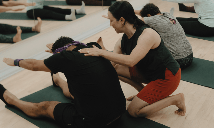 L'instructeur donne des corrections manuelles à un élève dans un cours de Pilates au sol en groupe dans un studio de St-Henri, Montréal, pendant que l'élève réalise l'exercice Spine Stretch Forward.