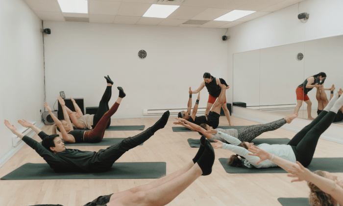 Un groupe d'élèves pratiquant l'exercice "Double Leg Stretch" sur des tapis de Pilates dans un studio de St-Henri, Montréal. Un élève est doucement corrigé et guidé par l'instructeur pour s'assurer de la bonne forme et de l'alignement.