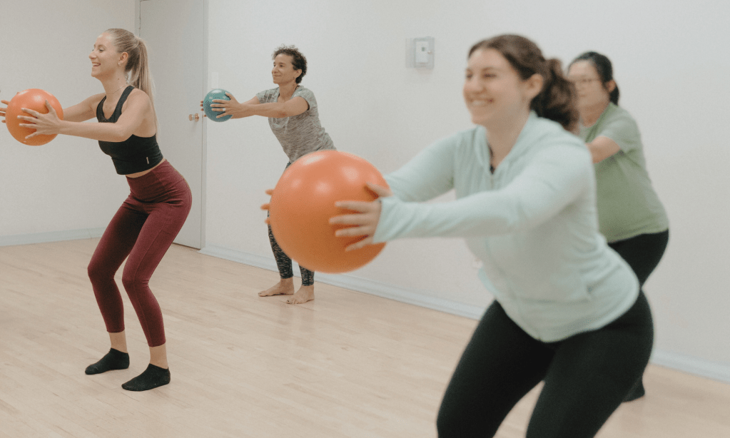 Étudiantes souriantes faisant un entraînement de barre à St-Henri, Montréal, utilisant un ballon de résistance pour ajouter un défi dans leur cours en groupe.