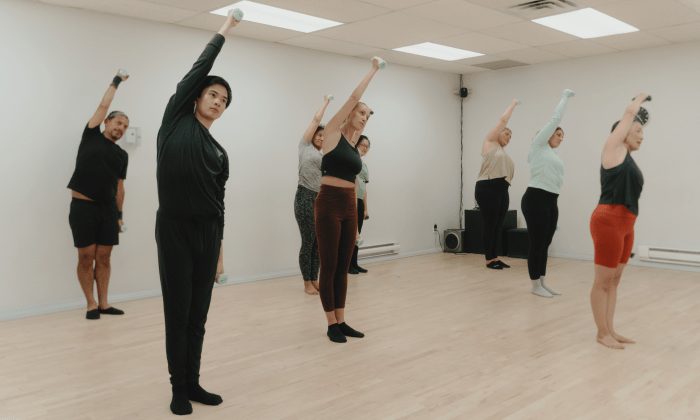Instructeur de barre fitness dirigeant un cours en groupe dans une flexion latérale debout avec des poids légers/haltères dans le cadre d'un entraînement de barre à St-Henri, Montréal.